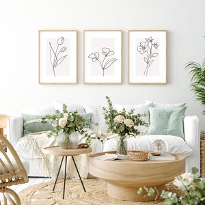 Può includere: Tre stampe minimaliste di arte lineare di fiori in bianco e nero, incorniciate in legno naturale, appese a una parete bianca sopra un divano bianco con cuscini verdi.