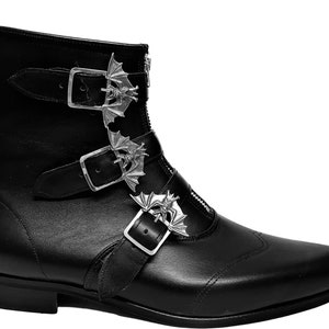 Może przedstawiać: Czarne skórzane botki z zaostrzonym noskiem i zamkiem błyskawicznym z boku. Botki mają trzy srebrne klamry z akcentami w kształcie nietoperza.