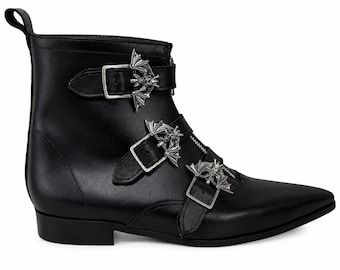 May include: A black ankle boot with a pointed toe and a low heel. The boot features a front zipper closure and three horizontal straps across the vamp, each secured with a silver-toned metal buckle shaped like a bat with outstretched wings.