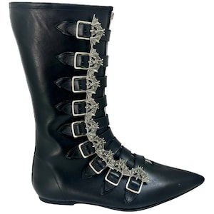 Może przedstawiać: Czarne skórzane kozaki za kolano ze spiczastym noskiem i wieloma srebrnymi klamrami w kształcie nietoperza.