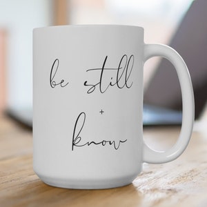 以下が含まれることがあります： 「be still + know」の黒い筆記体で書かれた白いセラミックマグ。マグカップには大きなハンドルと滑らかで光沢のある仕上げが施されています。コーヒー、紅茶、その他の飲み物に最適です。