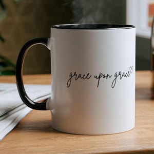 以下が含まれることがあります： 黒い縁と取っ手のある白いセラミック製のコーヒーカップ。カップには、小さなハートマークとともに、「grace upon grace」という文字が黒い筆記体で書かれています。