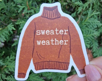 以下が含まれることがあります： 茶色のセーターの形をしたステッカーで、白い文字で「sweater weather」と書かれています。ステッカーは緑の葉の背景に表示されています。