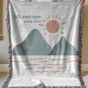 May include: A white woven throw blanket with a grey and blue mountain scene and a sun with the text "lift your eyes psalm 121:1" in black.