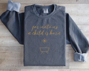 May include: A dark gray sweatshirt with the text "for unto us a child is born" in gold script. Below the text is a gold star and a manger symbol. The sleeves have light blue cuffs, and the words "more color options" are in the upper right corner.