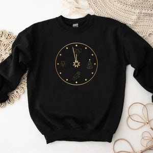 May include: Black sweatshirt with a gold clock design. The clock features a starburst at the center, with various celebratory icons around the clock face. The design is printed in gold on the black fabric. The sweatshirt is a casual, comfortable style.