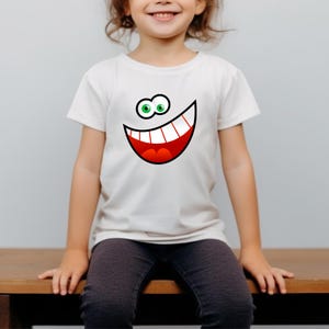 May include: A white t-shirt with a cartoon smiley face printed on the front. The face has large green eyes, a wide red mouth, and white teeth.
