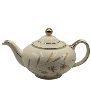 May include: A cream-colored teapot with a gold trim and floral design. The teapot has a lid with a gold knob.