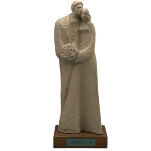 May include: A beige ceramic sculpture of a bride and groom standing together. The bride is holding a bouquet of flowers. The sculpture is on a wooden base with the words "Happily Ever After" inscribed on a plaque.