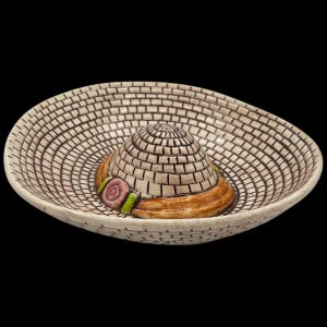 May include: A ceramic sombrero-shaped bowl with a brick-like pattern in beige and brown. The hat features a brown brim, a pink and green accent, and a conical crown. The bowl is designed with a wide, shallow brim.