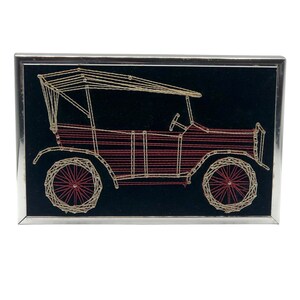 May include: A framed string art piece depicting a vintage car in red and gold thread on a black background.