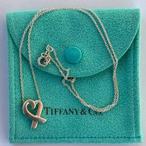 May include: A silver necklace with a heart and X pendant rests on a teal pouch. The pouch has a button closure and the words "TIFFANY & CO." printed on it. The necklace chain is delicate and the pendant is a polished silver.
