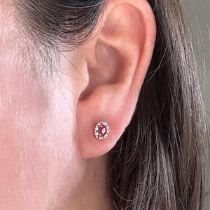 May include: A close-up of a small, oval-shaped stud earring. The earring features a central red gemstone surrounded by a halo of smaller, clear gemstones. The setting appears to be gold-toned, and the earring is worn on an ear.