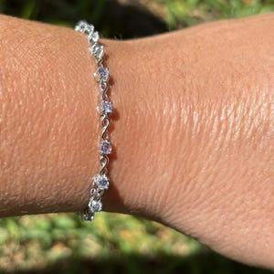 May include: A silver bracelet with a series of clear, round gemstones. The bracelet is delicate and appears to be made of a thin metal chain. The gemstones are evenly spaced along the chain, creating a sparkling effect.