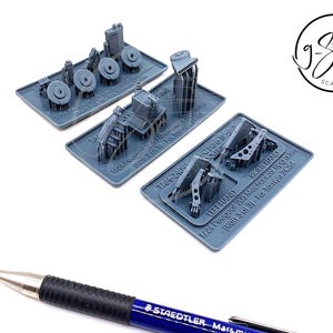 Op de afbeelding: Drie grijze plastic model auto onderdelen voor een Peugeot 205 op schaal 1:24. De onderdelen zijn gelabeld met "Linkerhand", "Rechterhand" en "TAM 24/6".