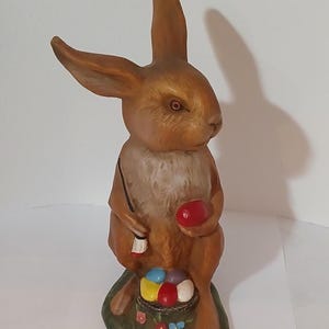 May include: A brown ceramic bunny figurine holding a red egg. The bunny is sitting on a green base with a basket of colorful eggs.