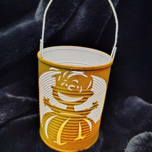 May include: A cylindrical metal can painted in a mustard yellow finish with a white wire handle. The exterior features a cutout design of a smiling cartoon character with outstretched arms, revealing the white interior of the metal container.