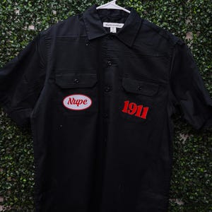 May include: Black button-up shirt with two chest pockets. Features a red and white oval patch that says "Nupe" and the number "1911" in red. The shirt is hanging on a white hanger against a green leafy background.