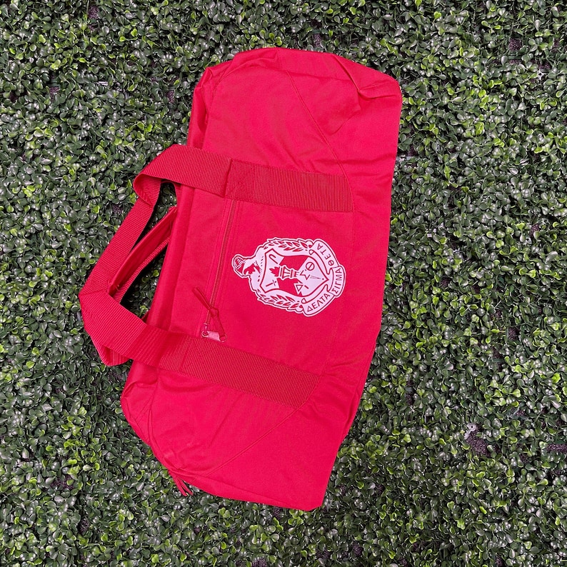 May include: A red duffel bag with a white embroidered crest featuring a crown, a key, and the text "Delta Sigma Theta".