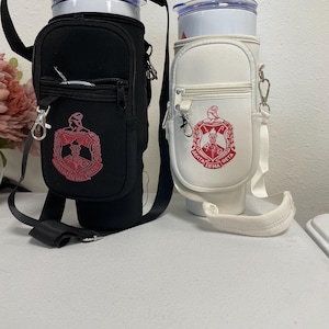 May include: Black and white insulated tumbler carriers with shoulder straps. The black carrier has a red emblem and zipper pocket. The white carrier has a red emblem and zipper pocket. Both carriers have a straw and are designed to hold a tumbler.
