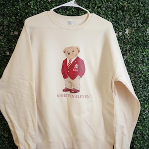 Puede incluir: Sudadera color crema con un oso de dibujos animados que lleva una blazer roja, pantalones color caqui y zapatos rojos. El oso tiene una corbata y una insignia que dice "KAW". El texto "NINETEEN ELEVEN" está impreso debajo.