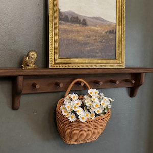 May include: A dark brown wooden wall-mounted coat rack with six pegs, featuring a gold-framed landscape painting above it, a small gold owl figurine on the left, and a woven wicker basket filled with white daisy flowers hanging from a peg.
