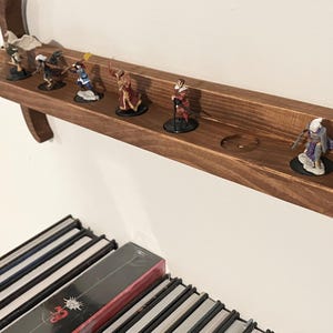 May include: A brown wooden wall-mounted display shelf shaped like a sword, featuring a long horizontal channel with circular indentations designed to hold small painted fantasy character figurines in a row along the blade section.
