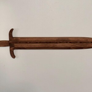 May include: A dark brown wooden wall-mounted display rack shaped like a sword. The blade section features a flat horizontal shelf with eight circular cutouts designed to hold items upright. The handle includes a circular pommel and a crossguard.