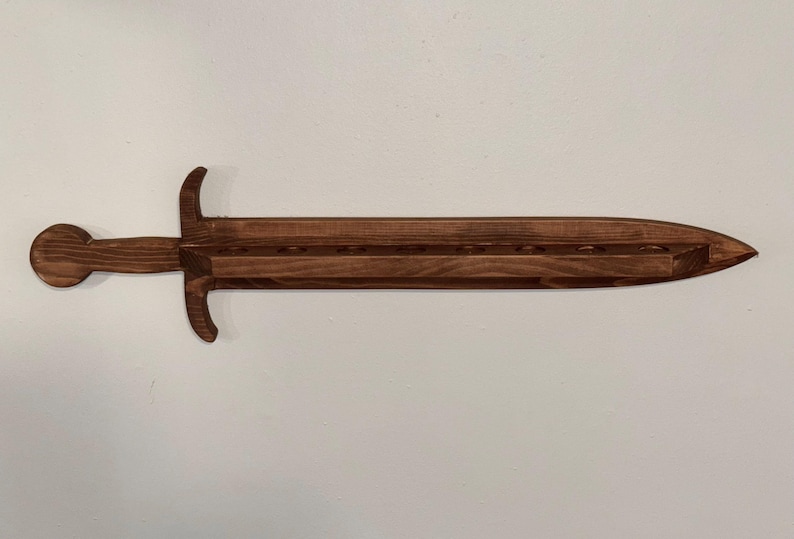 May include: A dark brown wooden wall-mounted display rack shaped like a sword. The blade section features a flat horizontal shelf with eight circular cutouts designed to hold items upright. The handle includes a circular pommel and a crossguard.
