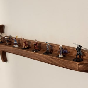 May include: A wall-mounted wooden shelf shaped like a longsword, featuring a dark brown wood grain finish. The shelf holds eight small, hand-painted fantasy character figurines standing in a row along the flat surface of the sword blade.