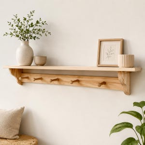 Könnte beinhalten: Ein Wandregal aus hellem Holz mit fünf Haken zum Aufhängen von Gegenständen. Auf dem Regal stehen eine gesprenkelte Keramikvase mit grünem Blattwerk, eine kleine Schale, ein gerahmtes Strichbild eines Blattes und ein gerippter zylindrischer Behälter.