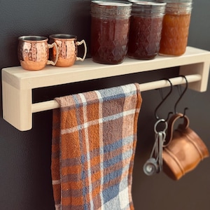 May include: A wooden shelf with a towel bar, holding copper mugs, jars of preserves, and a plaid towel. The shelf is made of light-colored wood and has a simple, modern design. The towel is orange, brown, and blue.
