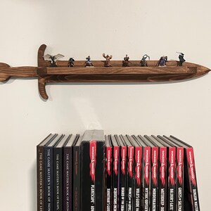 May include: A wall-mounted wooden shelf shaped like a sword with a dark brown finish. The shelf holds eight small fantasy character figurines. Below the shelf, a row of Dungeons & Dragons books is visible, including titles like Planescape and Xanathar's Guide.