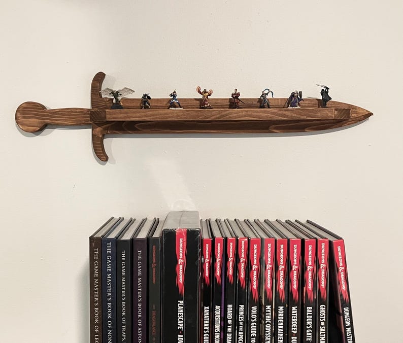 May include: A wall-mounted wooden shelf shaped like a sword with a dark brown finish. The shelf holds eight small fantasy character figurines. Below the shelf, a row of Dungeons & Dragons books is visible, including titles like Planescape and Xanathar's Guide.