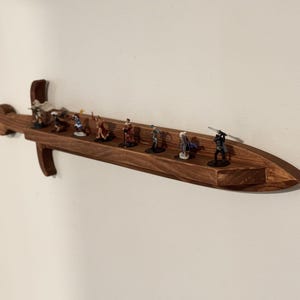 May include: A wall-mounted wooden display shelf shaped like a sword, featuring a long, flat surface for holding small items. The shelf is crafted from dark brown wood with a visible grain and holds eight small, painted character figurines.