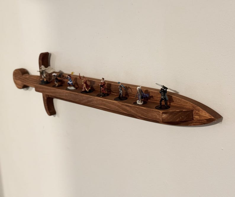 May include: A wall-mounted wooden display shelf shaped like a sword, featuring a long, flat surface for holding small items. The shelf is crafted from dark brown wood with a visible grain and holds eight small, painted character figurines.