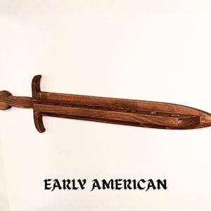 May include: A dark brown wooden wall-mounted display rack shaped like a sword. The blade section features a flat top surface with eight circular cutouts for holding items. The text "EARLY AMERICAN" is printed in black below the wooden sword.