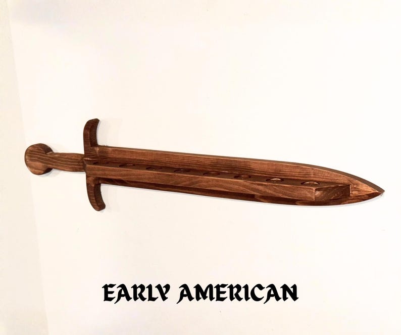 May include: A dark brown wooden wall-mounted display rack shaped like a sword. The blade section features a flat top surface with eight circular cutouts for holding items. The text "EARLY AMERICAN" is printed in black below the wooden sword.