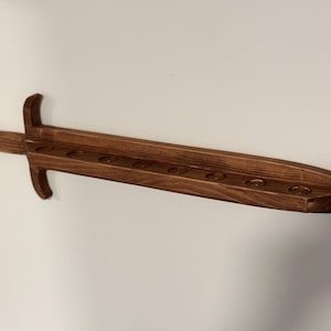 May include: A dark brown wooden wall-mounted display rack shaped like a sword. The blade section features a flat top surface with eight circular indentations aligned in a row, designed to hold small cylindrical objects securely in place.