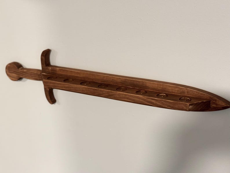 May include: A dark brown wooden wall-mounted display rack shaped like a sword. The blade section features a flat top surface with eight circular indentations aligned in a row, designed to hold small cylindrical objects securely in place.