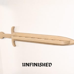 May include: A light-colored wooden sword-shaped wall shelf featuring a handle, a crossguard, and a blade with eight circular cutouts along the center. The word UNFINISHED is printed in black text below the wooden sword-shaped wall shelf.