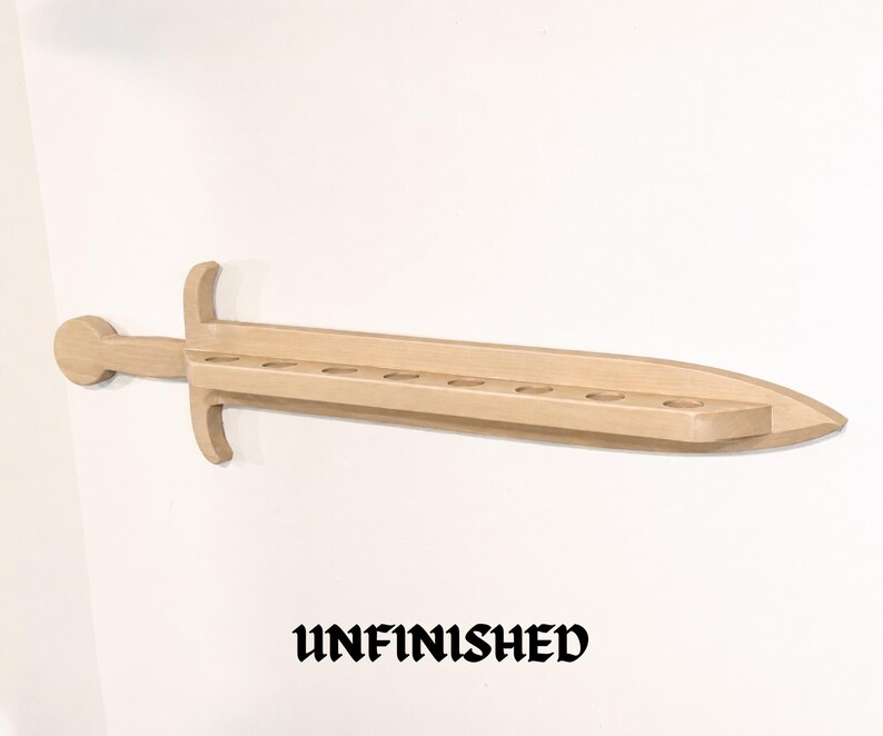 May include: A light-colored wooden sword-shaped wall shelf featuring a handle, a crossguard, and a blade with eight circular cutouts along the center. The word UNFINISHED is printed in black text below the wooden sword-shaped wall shelf.