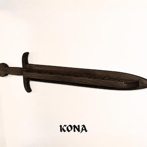 May include: A dark brown wooden wall-mounted shelf shaped like a sword, featuring a long blade with a series of circular cutouts along the center for holding items. The word KONA is printed in a bold, stylized black font below the sword shelf.