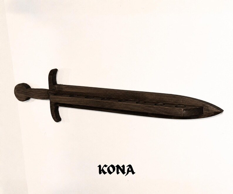 May include: A dark brown wooden wall-mounted shelf shaped like a sword, featuring a long blade with a series of circular cutouts along the center for holding items. The word KONA is printed in a bold, stylized black font below the sword shelf.
