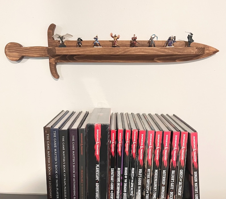 May include: A brown wooden wall shelf shaped like a sword, holding eight small fantasy character figurines. Below the shelf is a row of Dungeons & Dragons hardcover books, including titles like Planescape, Xanathar's Guide to Everything, and Ghosts of Saltmarsh.
