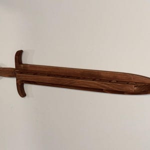 May include: A dark brown wooden wall-mounted display rack shaped like a sword. The horizontal blade section features a flat top surface with a series of circular cutouts designed to hold items, extending from a central hilt and a rounded pommel.