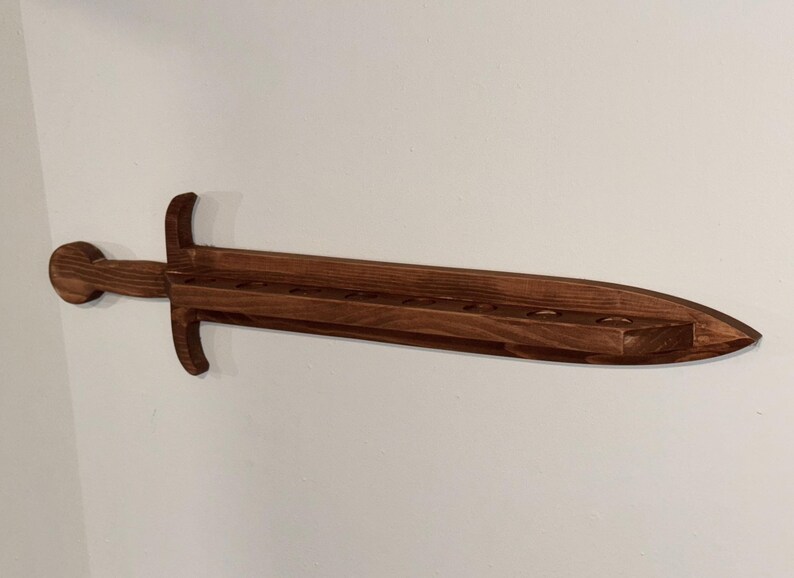 May include: A dark brown wooden wall-mounted display rack shaped like a sword. The horizontal blade section features a flat top surface with a series of circular cutouts designed to hold items, extending from a central hilt and a rounded pommel.
