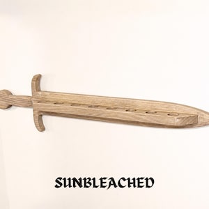May include: A light brown, sunbleached wooden sword-shaped wall shelf featuring a long, flat blade with eight circular cutouts along the top edge, a crossguard, and a rounded pommel. The word SUNBLEACHED is printed in black text below the object.