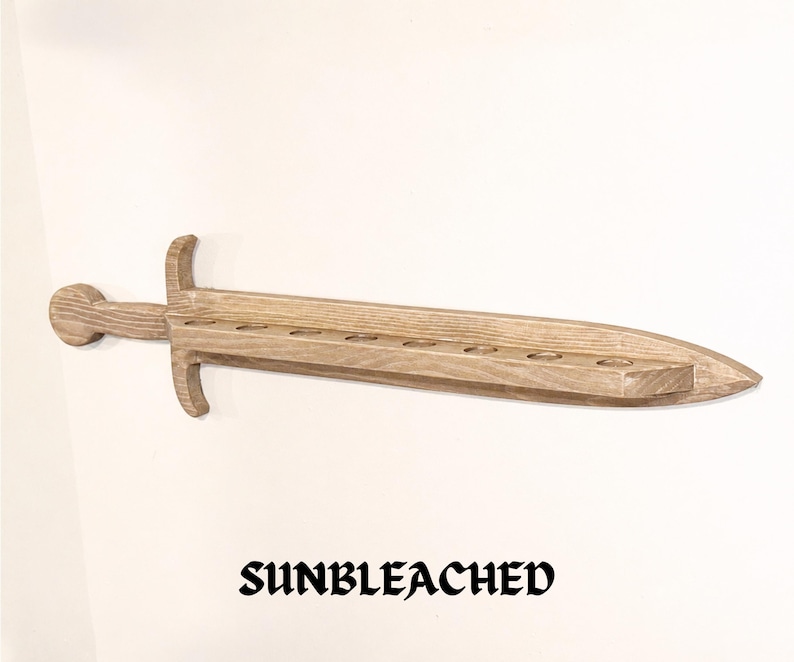 May include: A light brown, sunbleached wooden sword-shaped wall shelf featuring a long, flat blade with eight circular cutouts along the top edge, a crossguard, and a rounded pommel. The word SUNBLEACHED is printed in black text below the object.