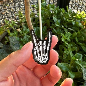 Pode incluir: Um alfinete de esmalte preto e branco em forma de m&atilde;o de esqueleto fazendo o gesto de rock and roll. O alfinete &eacute; segurado em uma m&atilde;o, com um fundo de folhagem verde e uma cerca de metal preta.